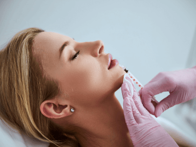 Woman lying with eyes closed as a clinician injects her chin during a cosmetic treatment with gloved hands visible.