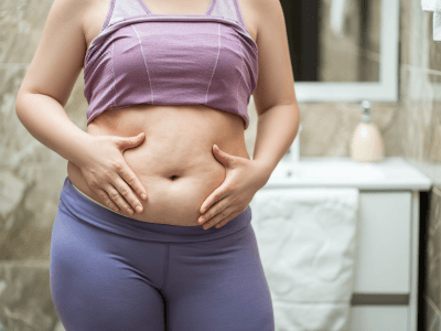 Tummy Tuck | Redefine Person wearing a purple sports bra and leggings pinching their belly in a bathroom.