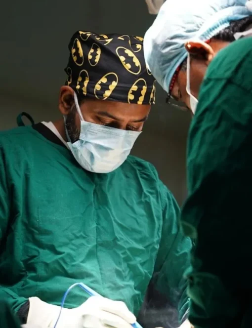 Surgeon in green scrubs and a Batman-pattern cap wearing a mask, focused on a procedure during surgery.