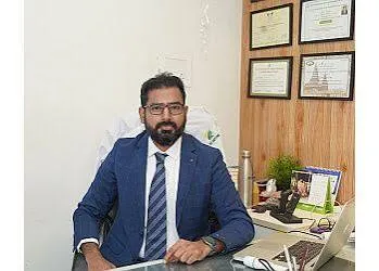 Man in a blue suit and striped tie sits at a desk in an office with framed certificates on the wall behind him.